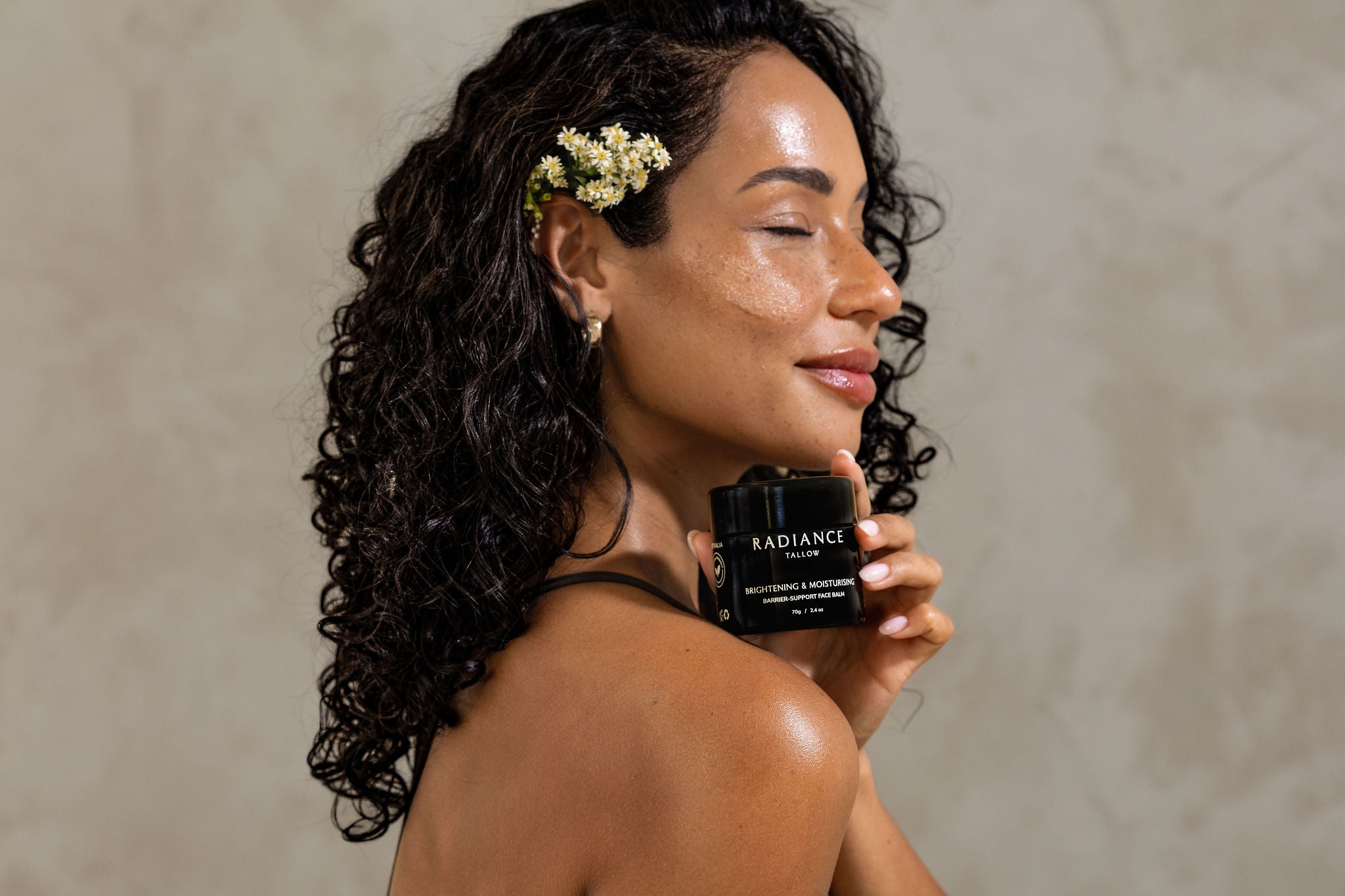 Woman holding a skincare product labeled 'Radiance tallow' against a neutral background and flowers in her hair