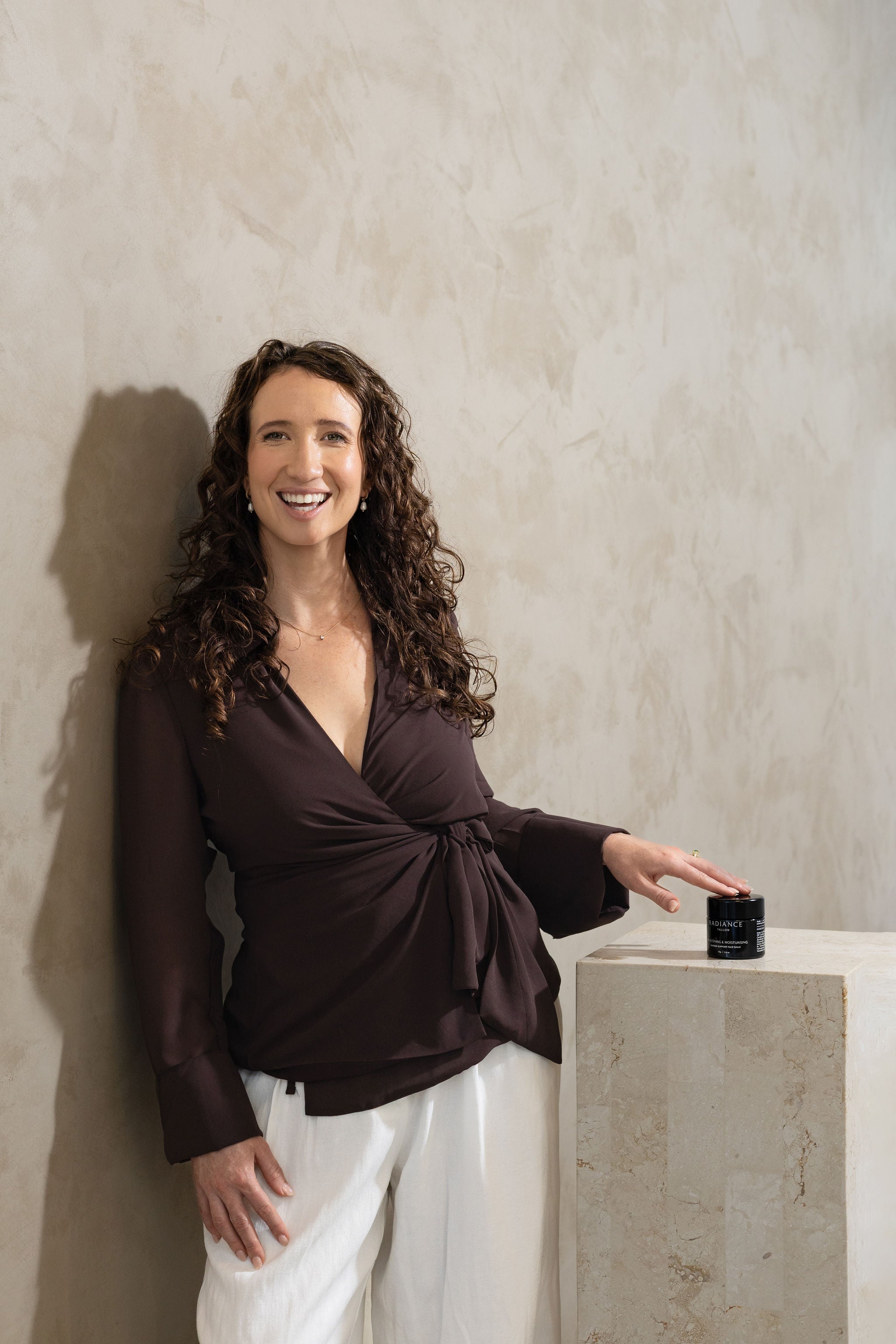 Woman , Emma Karst, in a brown top and white pants holding a small black container against a beige wall.